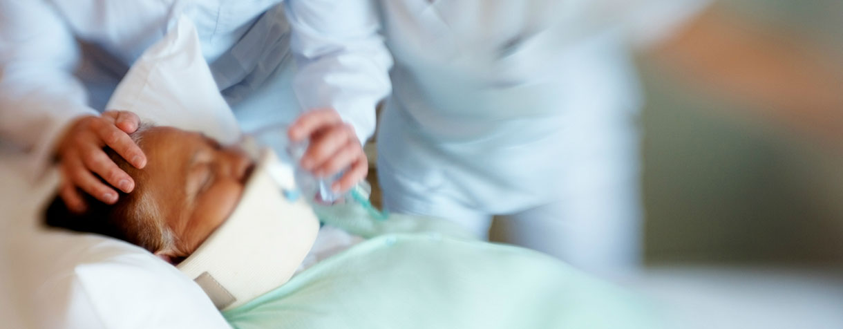 Two doctors standing behind a patient who has a neck brace. The doctor on the left is holding a breathing mask to the patient.